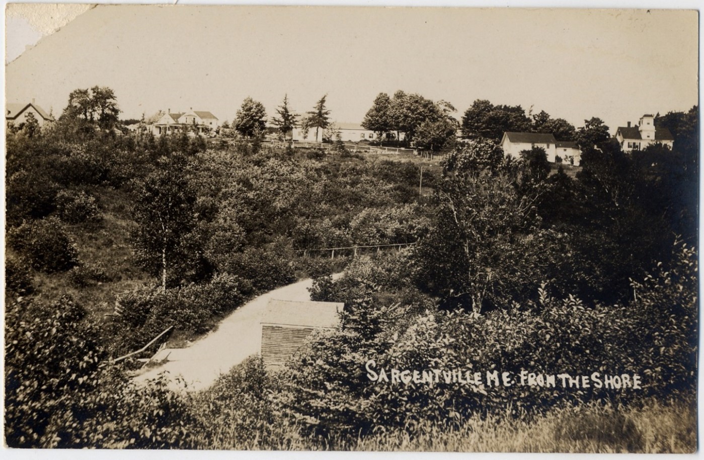A long-distance view of Rock Hill ( second house from the left) before Lydia Gower converted her barn to a residence. In the mid-foreground is the “powder house” on Shore Road where dynamite and other explosives were stored.